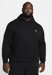 Μαύρο φούτερ με κουκούλα από μαλακό ύφασμα, με τσέπη καγκουρό, κουκούλα με κορδόνια και μικρό λογότυπο στο στήθος. Απλός, καθημερινός σχεδιασμός.