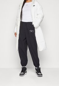 Vit puffjacka, svart åtsittande t-shirt med logotyp, svarta joggerbyxor med logotyp och svarta hög-top sneakers. Avslappnad atletisk outfit.