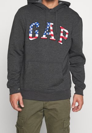 Hombre con sudadera con capucha color gris oscuro con letras "GAP" en patrón de la bandera estadounidense y pantalones cargo verde oliva, de pie contra un fondo liso.