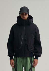 Schwarze gepolsterte Jacke mit hohem Kragen, mehreren Rei�ßverschluss-Taschen und passgenauem Bund. Getragen mit grünen Hosen und einer schwarzen Mütze.