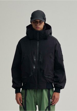 Schwarze gepolsterte Jacke mit hohem Kragen, mehreren Reißverschluss-Taschen und passgenauem Bund. Getragen mit grünen Hosen und einer schwarzen Mütze.