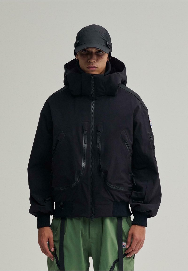 Schwarze gepolsterte Jacke mit hohem Kragen, mehreren Reißverschluss-Taschen und passgenauem Bund. Getragen mit grünen Hosen und einer schwarzen Mütze.