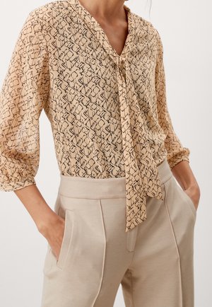 Frau trägt beige gemusterte Bluse mit vorderer Schnürung und hellbeige hoch taillierte Hose, Hände in den Taschen, gezeigt vom Hals bis zur Hüfte.
