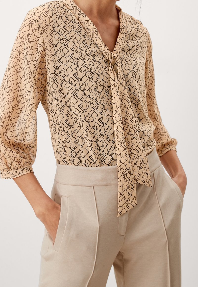 Femme portant un chemisier beige à motifs avec un nœud devant et un pantalon beige clair taille haute, mains dans les poches, montrée du cou jusqu'aux hanches.