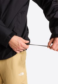Hænder justerer snoren i kanten af en sort jakke, som bæres sammen med beige North Face-bukser mod en ensfarvet hvid baggrund.