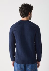Dunkelblauer Strickpullover mit gerippten Bündchen und Saum, der mit strukturierten Seitenpaneelen und Raglanärmeln ausgestattet ist, von hinten präsentiert.