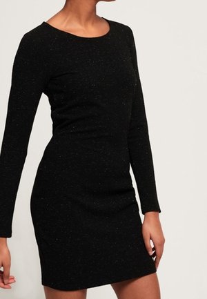 Woman wearing a fitted, long-sleeve, black dress with a round neckline against a plain light background.