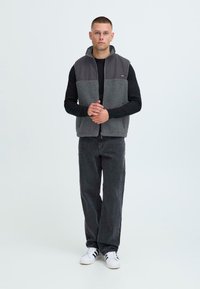 Grau Fleece-Weste mit einem dunkelgrauen oberen Abschnitt, getragen über einem schwarzen Langarmshirt; kombiniert mit dunklen Hosen und weißen Turnschuhen.