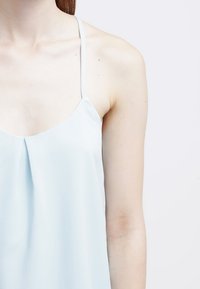 Light blue sleeveless top with thin straps; fabric is smooth with a slight sheen, featuring a pleated detail at the front.