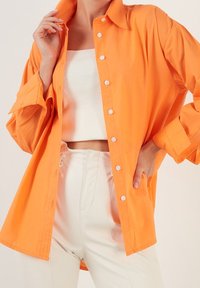 Orange stor skjorta med knappar och långa ärmar, gjord av lätt tyg. Bärs över en vit ribbad croptop och kombineras med vita byxor.