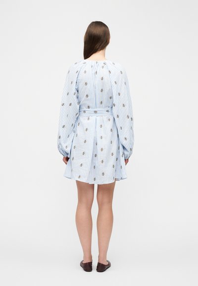 Γυναίκα με την πλάτη, φοράει μπλε και άσπρο ριγέ φόρεμα με σχέδια μέλισσας, με φουσκωτά μακριά μανίκια, ζώνη στη μέση και καφέ φλατ παπούτσια.