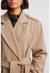 Beige visgraat jas met grote gesneden revers, wijde mouwen en een strikceintuur. Heeft een voorzak en knoopaccenten op de schouders.