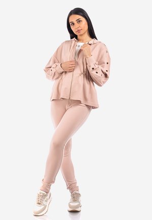 Giacca a zip rosa con cappuccio e coulisse, abbinata a leggings coordinati. Texture liscia e dettagli in oro sulle rifiniture. Sneakers bianche completano l'outfit.