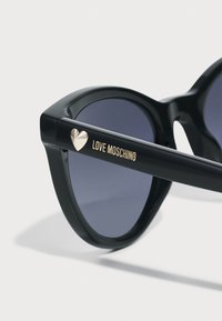 Juodos katės akių stiliaus saulės akiniai su tamsiais lęšiais, turintys auksinį širdies akcentą ir užrašą "LOVE MOSCHINO" ant rankos. Lygus plastikinis paviršius.