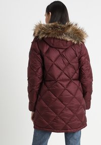 Bordeaux pufferjakke med diamantquiltet mønster, pelsforet hætte og en glat tekstur, båret over jeans i blå.