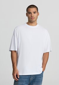 Mężczyzna z krótkimi włosami nosi gładką, dużą białą koszulkę i niebieskie jeansy, stojąc na tle jasnoszarym.