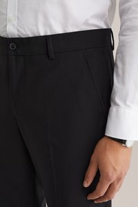 Pantalon noir ajusté en tissu lisse, avec des poches avant et une passante de ceinture. La tenue est associée à une chemise blanche à boutons et une montre au poignet.