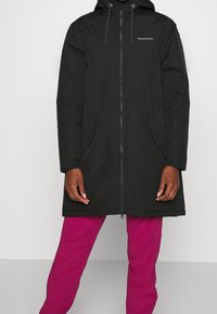 Zwarte waterdichte lange jas met capuchon, voorzien van een rits aan de voorkant, zijzakken en een gladde, strakke textuur. Draagt over roze broek.