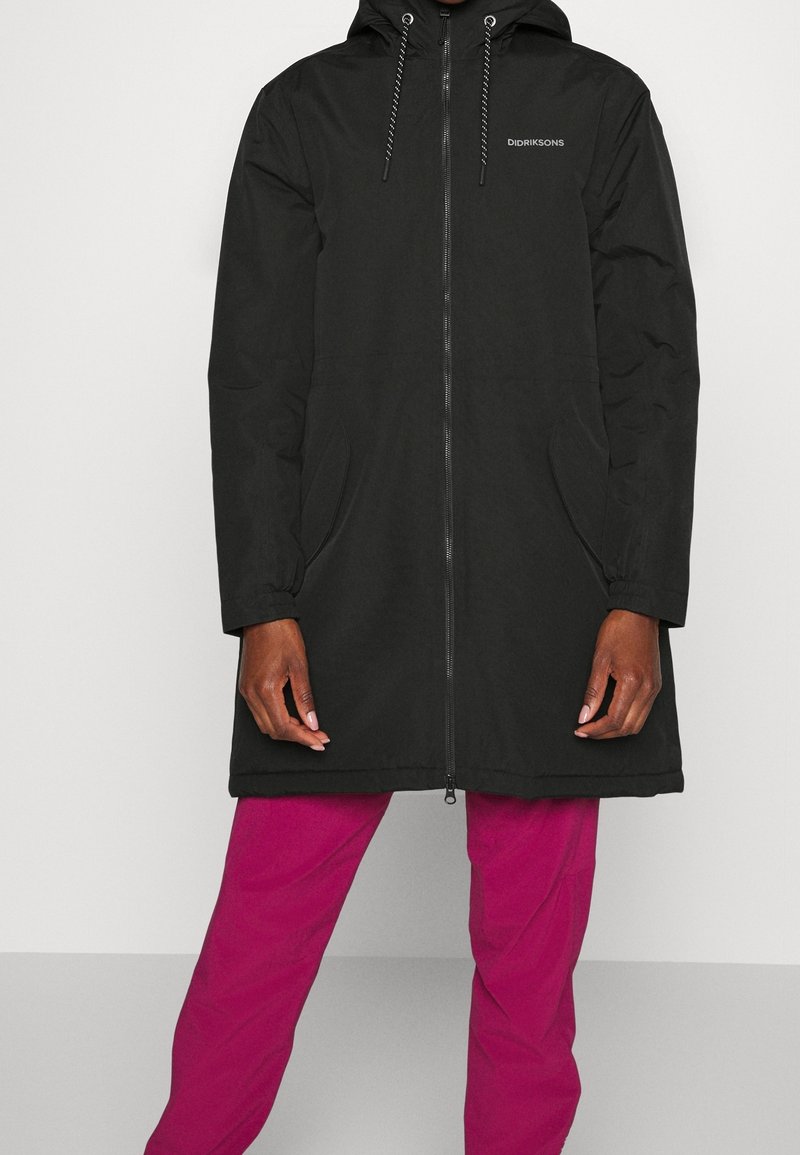Zwarte waterdichte lange jas met capuchon, voorzien van een rits aan de voorkant, zijzakken en een gladde, strakke textuur. Draagt over roze broek.