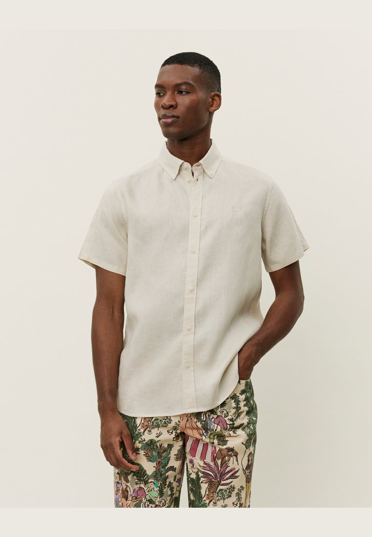 Camisa corta beige claro con frente abotonado y cuello vuelto; llevada con pantalones verdes y beige de patrón que presentan diseños florales.
