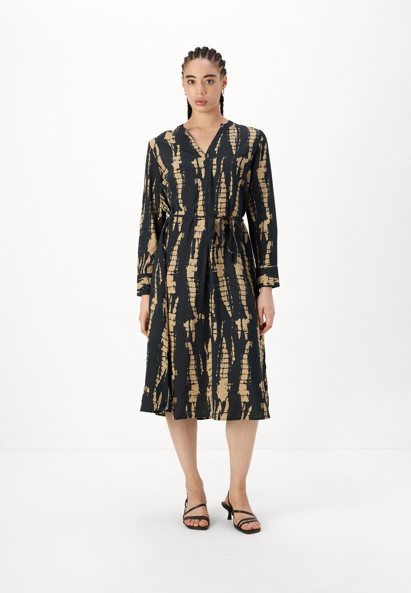 Juoda ir auksinė raštuota suknelė su ilgomis rankovėmis ir juosmens dirželiu, turinti V formos iškirptę ir plaukuojančią iki kelių sijoną. Minimalistinis dizainas.