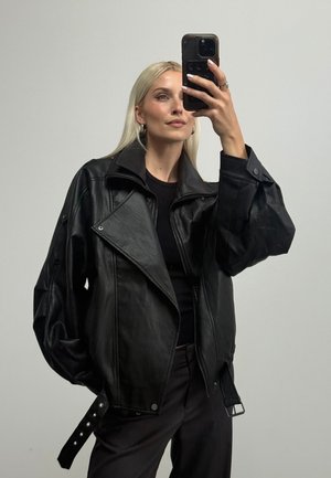 Blonde Frau trägt eine schwarze, übergroße Lederjacke und schwarze Hose und macht ein Selfie mit einem Smartphone vor einem einfarbigen Hintergrund.