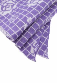 Plaid in pile viola con un motivo a griglia bianco, caratterizzata da una texture morbida e bordi frangiati. Piegata con un dettaglio di design visibile.