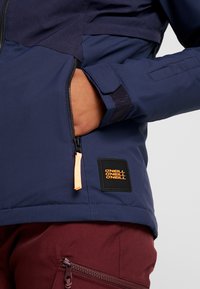 Giacca blu navy con tasca con zip e accenti arancioni. Presenta un logo in tessuto testurizzato. Indossata sopra pantaloni color bordeaux.