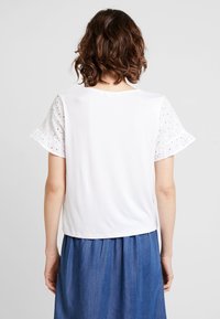 Haut blanc avec des manches courtes volantées en dentelle, doté d'un col rond et d'un ourlet légèrement courbé ; porté sur une jupe en denim.