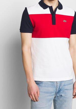 Mand iført en poloshirt i farveblok med marineblå ærmer og krave, rød bryststribe, hvid top og bund, kombineret med lyseblå jeans.