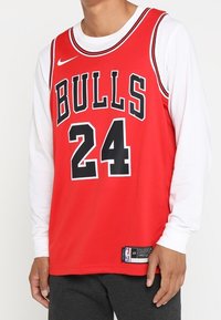 Červený basketbalový dres s nápisem "BULLS" v černé barvě na přední straně, číslem "24" pod ním a logem Nike na hrudi. Přes dres je vrstvená bílá košile s dlouhým rukávem.