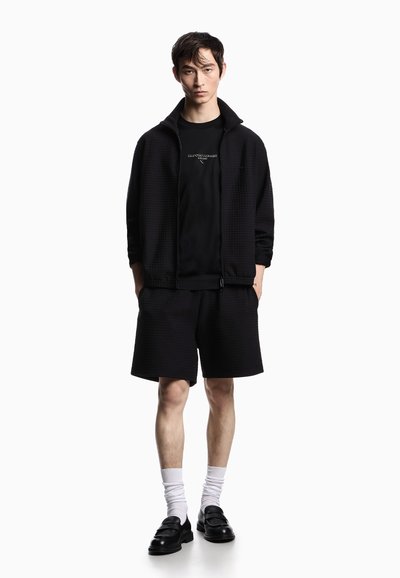 Uomo che indossa una giacca nera a zip con texture, pantaloni corti abbinati, maglietta nera con logo bianco, calzini bianchi e mocassini neri, in piedi davanti a uno sfondo bianco.