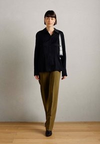 Camicia nera con bottoni e maniche lunghe, abbinata a pantaloni verde oliva. Scarpe nere e una borsa a tracolla nera completano l'outfit.
