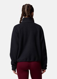 Persoon met twee gevlochten paardenstaarten, draagend een zwart fleecejack en donkerrode legging, met de rug naar voren tegen een effen achtergrond.