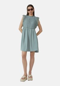 Robe bleu clair avec un haut en dentelle et des manches volantées. Jupe fluide, associée à des sandales compensées crème et des lunettes de soleil marron.