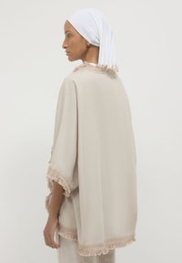 Donna con una sciarpa frangiata beige sopra un abbigliamento chiaro e un velo bianco, in piedi di profilo su uno sfondo semplice.