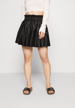 Black faux leather skirt with a pleated design and an elastic waistband. Worn with a light-colored sweater and flat black sandals.