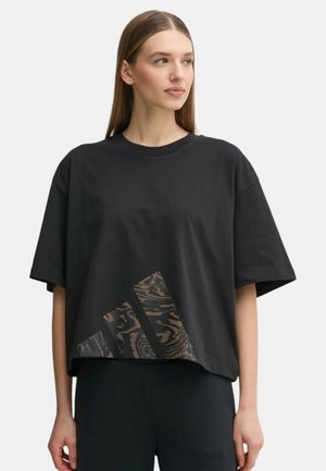 Donna che indossa una camicia nera a maniche corte larga con un motivo triangolare astratto marrone sul davanti, abbinata a pantaloni neri.