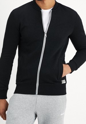 Chaqueta negra con cremallera y cuello redondo, con una cremallera gris y un bolsillo en el costado. Lleva puesta una camiseta blanca y pantalones grises.