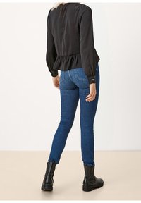 Top peplum nero con maniche lunghe, abbinato a jeans skinny blu e stivaletti neri, visto di spalle. Dettaglio semplice di chiusura con bottone.