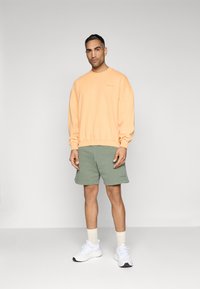 Felpa over-sized color pesco e pantaloni corti verde oliva, abbinati a sneakers bianche e calzini bianchi. La felpa presenta un sottile dettaglio di testo.