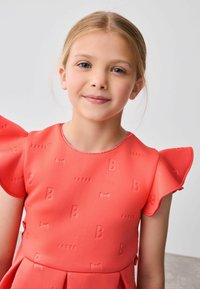 Koralinė suknelė su iškiliais raštais - lankeliais ir raidėmis "B" bei "BAKER." Turi trumpos ruffles rankoves ir plačią sijoną. Lygi tekstūra.