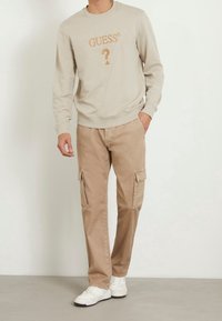 Sudadera color beige claro con el logo "GUESS" y un gráfico de signo de interrogación. Combinada con pantalones cargo color caqui que tienen bolsillos laterales. Zapatillas blancas.