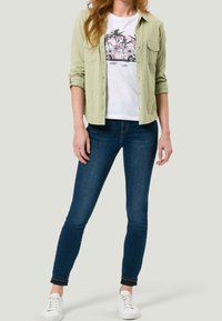 Camicia button-up verde chiaro sopra una t-shirt bianca con una grafica di una macchina vintage rosa, abbinata a jeans skinny blu scuro e sneakers bianche.