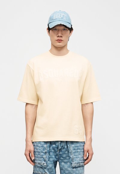 Νεαρός άνδρας που φοράει ανοιχτό μπεζ φαρδιά μπλούζα με ανάγλυφο κείμενο και αριθμό, μπλε τζιν σορτς με σχέδιο και ανοιχτό μπλε καπέλο με τη λέξη "HOT".