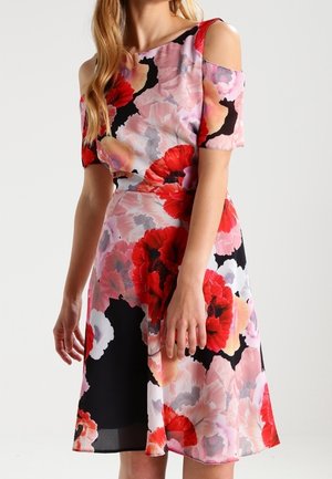 Mujer vistiendo un vestido de hombros descubiertos con grandes patrones florales rojos, rosados y negros sobre un fondo claro.