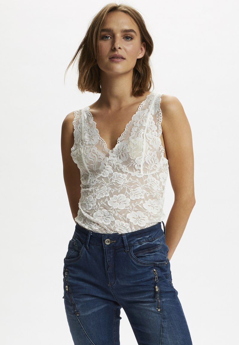 Weißes Spitzen-Bodysuit mit floralen Mustern, tiefem V-Ausschnitt und gewellten Rändern, kombiniert mit dunklen Jeans aus Denim mit Reißverschlussdetails.