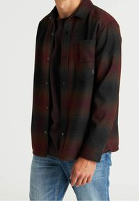 Camicia di flanella a quadri rossa e nera con chiusura frontale a bottoni, una tasca al petto e una vestibilità leggermente ampia. Indossata sopra una t-shirt scura, abbinata a un paio di jeans.