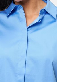 Chemise bleu clair à boutons avec col pointu, en tissu lisse, présentant une couture frontale et un éclat subtil.