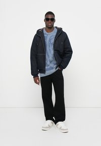 Jeans jakna s kapuco, temno modra, z dvema sprednjima žepoma, v kombinaciji s svetlomodrim puloverjem z vzorcem, črnimi hlačami in belimi športnimi copati.